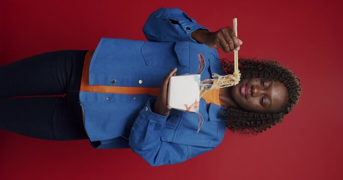 Vertical video of African american woman looking at camera and smiling, preparing to eat noodles, seafood and vegetables with chopsticks on isolated red background. Asia street food.