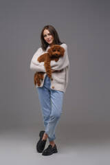 Stylish young brunette woman in casual wear cradling her cherished brown toy poodle, posed before a...