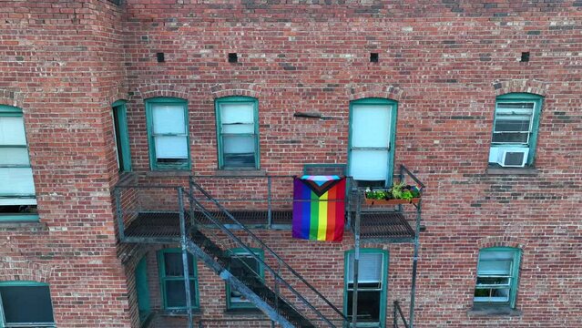 Gay pride and transgender ally flag on fire escape of old brick inner city apartment building. LGBTQIA+ theme. Aerial push in.