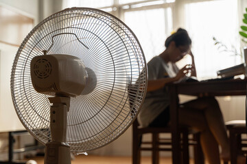 Asian unhappy woman sitting in front of working fan suffering from heat in modern house on sunny summer day.