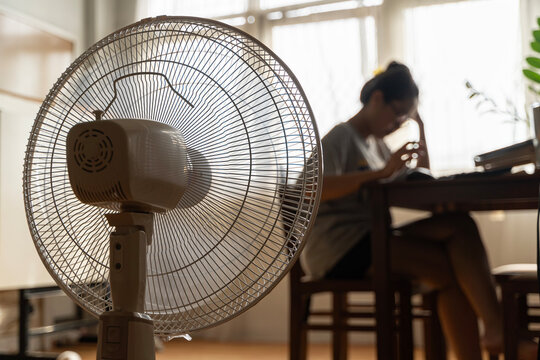 Asian Unhappy Woman Sitting In Front Of Working Fan Suffering From Heat In Modern House On Sunny Summer Day.