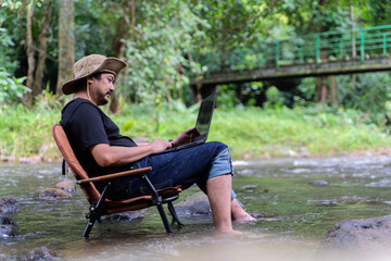 Fototapeta premium Handsome bearded Asian man of working age sitting on camping chair and working on laptop near stream in middle of park in outdoor