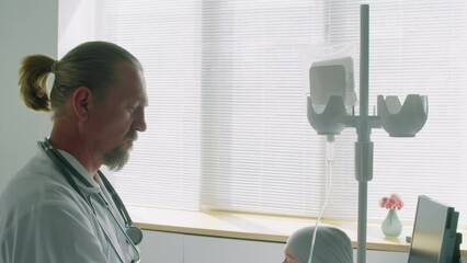 Tilt down shot of male doctor talking to female patient with cancer laying with dropper for chemotherapy in ward