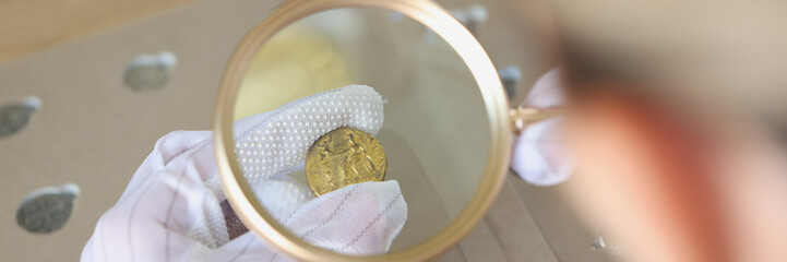 Gloved specialist examining gold coin with magnifying glass.