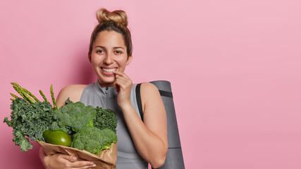 Sport and health. Indoor photo of young pretty happy smiling European lady standing on left isolated on pink background wearing grey tracksuit holding bag with vegetables with space for promotion