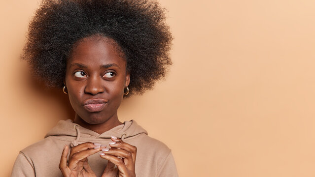 People and emotions. Indoor photo of young happy African american woman standing on left isolated on beige background wearing hoodie keeping hands raised with blank space for your advertisement