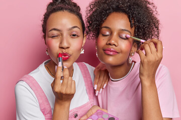 Photo of female friends apply red lipstick and eyeshadow put on makeup use cosmetic brush stand next to each other preapre for party isolated over pink background. Beauty and self care concept - obrazy, fototapety, plakaty