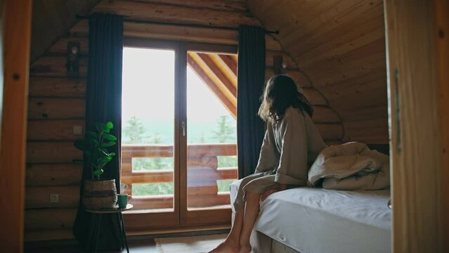 A young woman in a bathrobe is sitting on the bed and enjoying the view from the mountain chalet window after waking up.