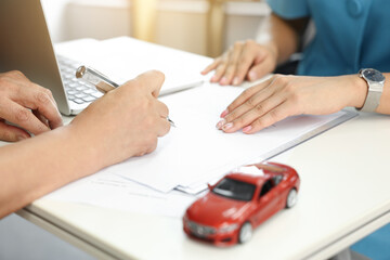 자동차 모형과 서류가 놓여진 데스크 위에서 자동차보험 계약에 서명하는 손  - obrazy, fototapety, plakaty