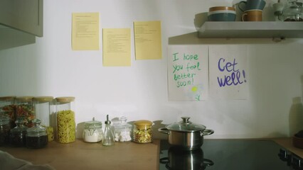 Medium shot of kitchen wall with recovery wishes, stove with pan and other utensil