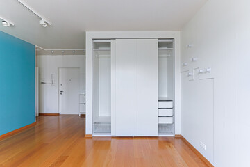 interior of an empty apartment with white walls and wooden floor, open closet, dressing room with storage system, bright blue wall