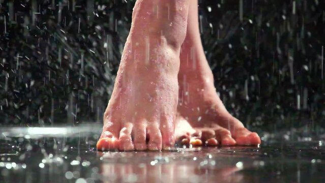 Barefoot woman with crossed legs stands on tiptoes in rain. Female with neat nails stands on mirror surface slow motion