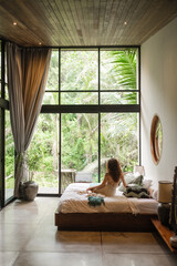 A girl of European appearance, with curly hair, greets the morning in a luxurious villa on the bed, admiring the nature outside the large window. - obrazy, fototapety, plakaty