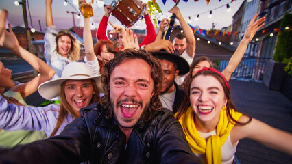 Young caucasian man making video with his friends on a party using smartphone. Rooftop party friends having fun drinking beer and cocktails.