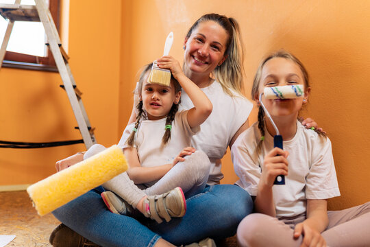 young mother and her daughters are sitting on the floor of the room with brushes and rollers in front of the wall, having fun
