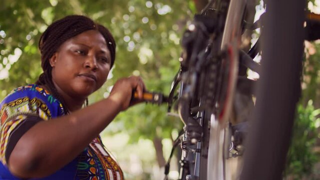 Dedicated sporty african american woman checking performance of bicycle parts by spinning the pedals. Active female cyclist ensuring proper function of tire close-up of bike wheel in home yard.
