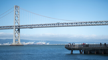 A fishing pier extends into the San Francisco Bay. In the background, the Bay Bridge, connecting San Francisco with East Bay / Oakland, towers over the scene.