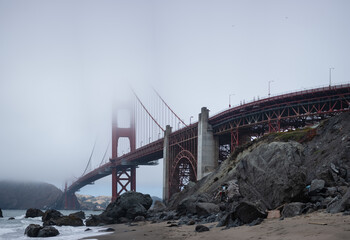 The Golden Gate Bridge looms tall over an empty, rocky beach on a foggy day. In the foreground,...