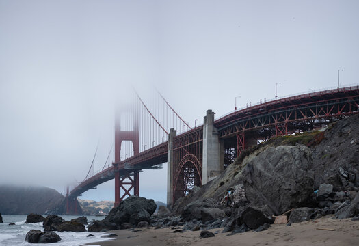 The Golden Gate Bridge looms tall over an empty, rocky beach on a foggy day. In the foreground, massive boulders are littered across the sand. - Powered by Adobe