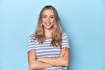 Blonde young caucasian woman in blue studio who feels confident, crossing arms with determination.