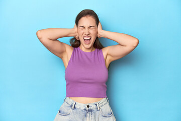 Fashionable young woman in a purple top on blue background covering ears with hands trying not to hear too loud sound.