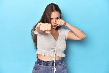 Stylish young woman in white blouse on a blue studio backdrop throwing a punch, anger, fighting due...
