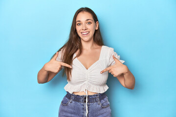 Stylish young woman in white blouse on a blue studio backdrop surprised pointing with finger,...