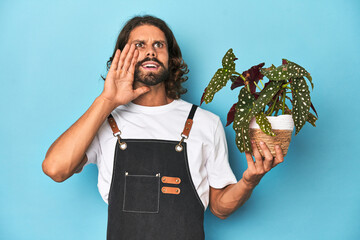 Long-haired gardener with beard holding a plant shouting and holding palm near opened mouth.