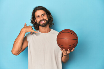 Bearded man with basketball in blue studio showing a mobile phone call gesture with fingers.