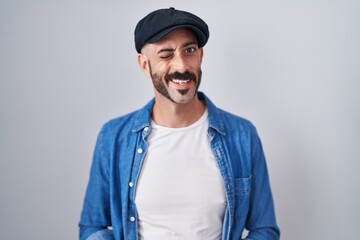 Hispanic man with beard standing over isolated background winking looking at the camera with sexy expression, cheerful and happy face.
