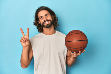Bearded man with basketball in blue studio joyful and carefree showing a peace symbol with fingers.