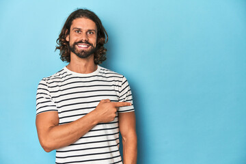 Bearded man in a striped shirt, blue backdrop smiling and pointing aside, showing something at blank space.