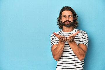Bearded man in a striped shirt, blue backdrop folding lips and holding palms to send air kiss.
