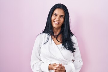 Mature hispanic woman standing over pink background with hands together and crossed fingers smiling relaxed and cheerful. success and optimistic