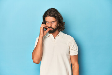 Long-haired man in a white polo, blue studio with fingers on lips keeping a secret.