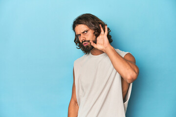 Bearded man in a tank top, blue backdrop trying to listening a gossip.