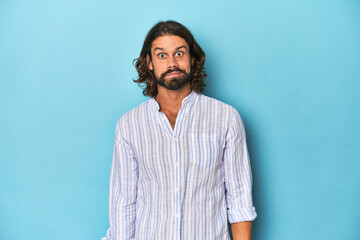 Man with beard in blue striped shirt, blue studio shrugs shoulders and open eyes confused.