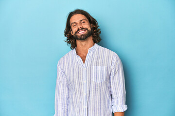Man with beard in blue striped shirt, blue studio laughs and closes eyes, feels relaxed and happy.