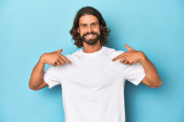 Bearded man in a white shirt, blue backdrop surprised pointing with finger, smiling broadly.