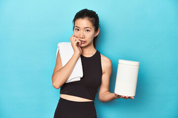Young Asian sportswoman with protein bottle, gym setup, biting fingernails, nervous and very anxious.