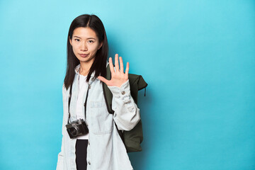 Young Asian traveler ready to capture adventures, smiling cheerful showing number five with fingers.