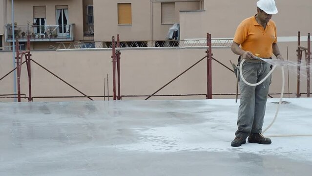 Construction worker watering fresh concrete slab using a hose.