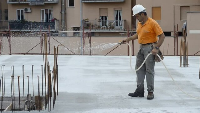 Construction worker watering fresh concrete slab using a hose.