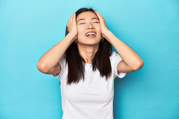 Young Asian woman in white t-shirt, studio shot, laughs joyfully keeping hands on head. Happiness concept.