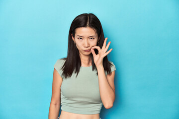 Asian woman in summer green top, studio backdrop, with fingers on lips keeping a secret.