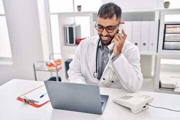 Fototapeta na wymiar Young hispanic man doctor using laptop talking on telephone at clinic