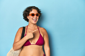 Young woman in bikini with beach bag smiling and pointing aside, showing something at blank space.