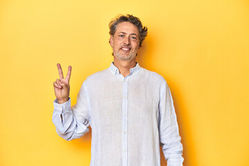 Middle-aged man posing on a yellow backdrop showing number two with fingers.