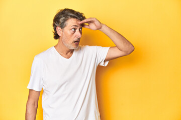 Middle-aged man posing on a yellow backdrop looking far away keeping hand on forehead.