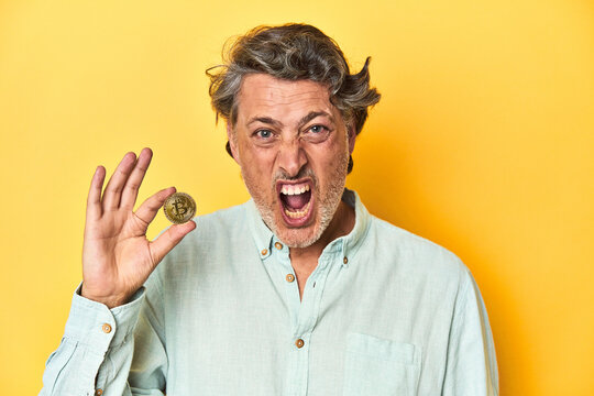 Man holding a physical bitcoin, yellow backdrop screaming very angry and aggressive.